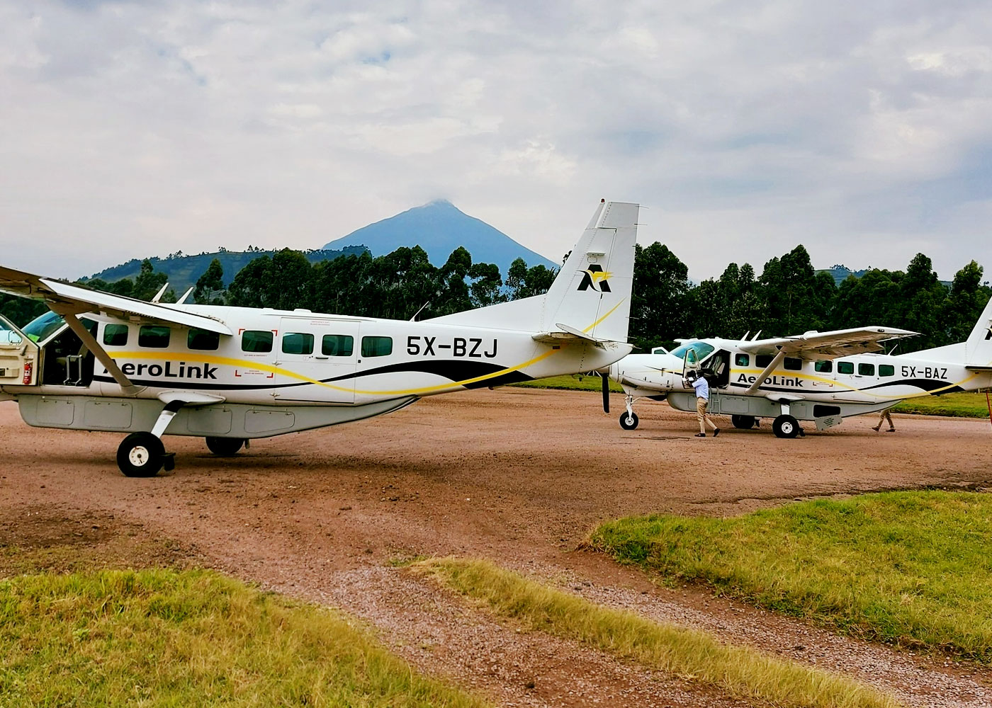Two small AeroLink airplanes are parked on a dirt airstrip, ready for a 3 Days Bwindi Gorilla Habituation Fly in Experience, with trees and a mountain visible in the background under a cloudy sky.